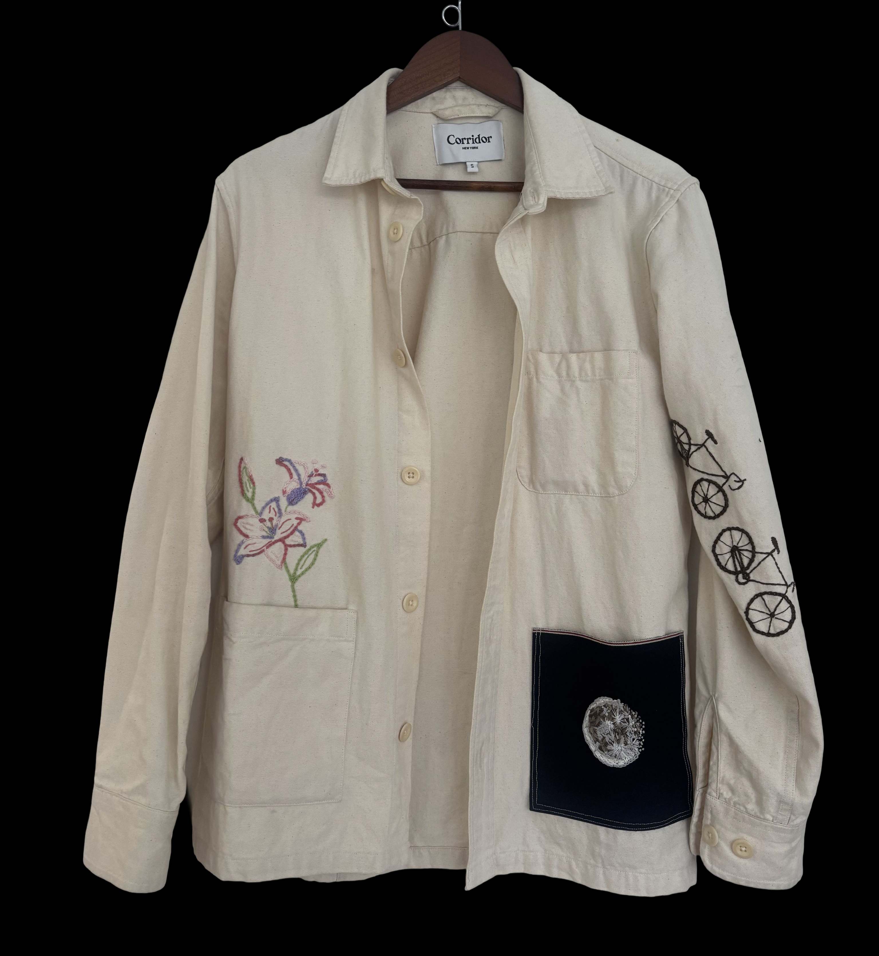 Natural cotton chore jacket with embroidered lilies, a bicycle down the sleeve, and a navy patch